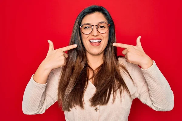赤い隔離された背景の上に立つメガネを身に着けている若いヒスパニック系のスマートな女性は 明るい表情と指の歯と口で指して笑っています 歯科衛生の概念 — ストック写真