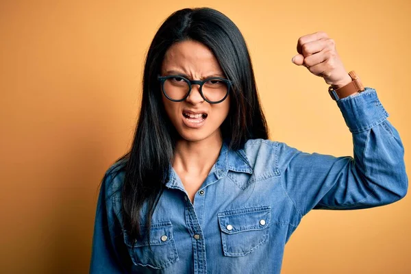 年轻美丽的中国女人穿着休闲裤衬衫 披着孤立的黄色背景 怒气冲冲地举起拳头 怒气冲冲地大叫着 愤怒和好斗的概念 — 图库照片