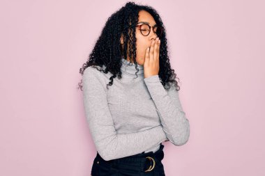 Genç Afro-Amerikalı kadın balıkçı yaka kazak ve pembe arka plan üzerine gözlük takmış esnemekten sıkılmış, ağzı eliyle kapatmaktan sıkılmış. Huzursuz ve uykulu..