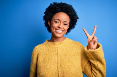Kıvırcık saçlı, sarı kazaklı, genç, Afro-Amerikan bir kadın. İki numarayı gösterip, parmaklarını gösterirken, kendinden emin ve mutlu gülümsüyor..