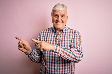 Üst düzey yakışıklı, parlak gömlekli, soyutlanmış pembe arka planda gülümseyen ve kameraya bakan, iki eliyle ve parmaklarıyla kenarı işaret eden bir adam..