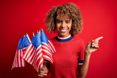 Genç Afro-Amerikan vatansever kıvırcık kadın kırmızı arka planda Birleşik Devletler bayraklarını tutuyor.