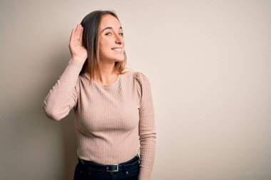 İzole edilmiş beyaz arka planda dikilen sıradan bir süveter giyen genç, güzel bir kadın. Kulağı kulağında, dedikoduları ve dedikoduları dinliyor. Sağırlık kavramı.