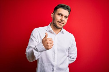 Mavi gözlü, zarif gömlekli, kırmızı izole edilmiş arka planda duran genç bir iş adamı mutlu bir el hareketi yapıyor. Kameraya bakıp başarıyı gösteren ifade onaylanıyor.