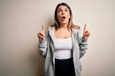 Genç ve güzel esmer bayan sporcu kadın beyaz arka plan üzerinde spor giyiniyor hayret ve şaşkınlık içinde yukarı bakıyor ve parmaklarıyla işaret ediyor ve kollarını kaldırıyor..