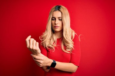 Genç, sarışın, gündelik tişört giyen, izole edilmiş kırmızı arka planda duran, ellerinde ve parmaklarında ağrı çeken, iltihaplı, eklem iltihabı olan bir kadın.