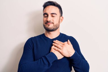 Sakallı, sıradan bir süveter giyen, göğsünde elleri göğsünde, yüzünde minnet dolu bir jest ile gülümseyen genç yakışıklı bir adam. Sağlık kavramı.