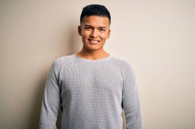 Yüzünde mutlu ve havalı bir gülümsemeyle izole edilmiş beyaz arka planda dikilen gündelik süveter giyen genç, yakışıklı latin adam. Şanslı kişi..