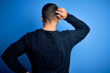 İzole edilmiş mavi arka plan üzerinde duran sıradan bir süveter giyen genç yakışıklı adam geriye dönüp, elleri kafasında şüpheyi düşünüyor.