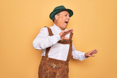 Kıdemli gri saçlı adam sarı arka planda geleneksel Alman Ekim Festivali kıyafeti giyiyor mutlu ve neşeli dans ediyor, hareketli ve kendinden emin bir şekilde müzik dinliyor.