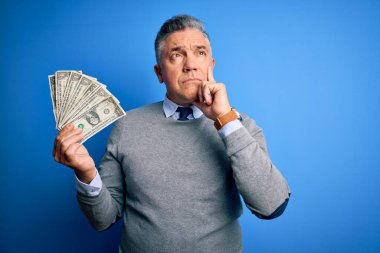 Orta yaşlı, yakışıklı, gri saçlı, elinde bir sürü dolar tutan, mavi arka planda ciddi bir yüzü olan, soru soran, kafası karışık bir adam.
