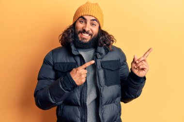 Genç Arap adam kışlık kıyafetlerini giyiyor gülümsüyor ve kameraya bakıyor iki eliyle ve parmaklarıyla kenarı işaret ediyor.. 