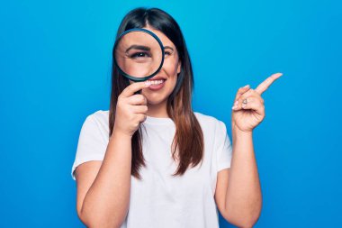 Genç, güzel esmer kadın, izole edilmiş mavi arka planda büyüteç kullanıyor. Gülümsüyor, mutlu bir şekilde elini ve parmağını yana doğru gösteriyor.