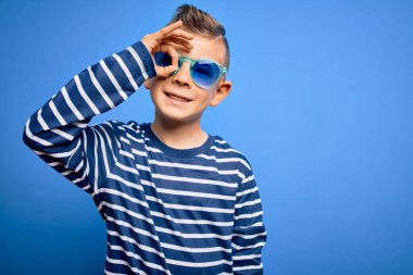 Mavi gözlü küçük beyaz çocuk mavi arka planda güneş gözlüğü takıyor. El hareketleriyle gülümsüyor ve mutlu yüzüyle parmaklarının arasından bakıyor..