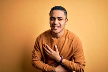 İzole edilmiş sarı arka planda dikilen sıradan bir süveter giyen genç Brezilyalı adam. Gülerek kahkahalarla gülüyor..