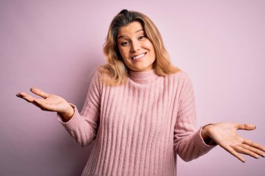 İzole edilmiş arka planda pembe süveter giyen genç, güzel, sarışın bir kadın. Kolları ve elleri havada, kafası karışmış bir ifade. Şüphe kavramı.