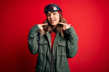 Kırmızı zemin üzerinde kayak havası için kayak gözlüğü ve kışlık palto giyen genç sarışın kız gülümseyip, parmaklarını ve ağzını işaret ediyor. Diş sağlığı konsepti.