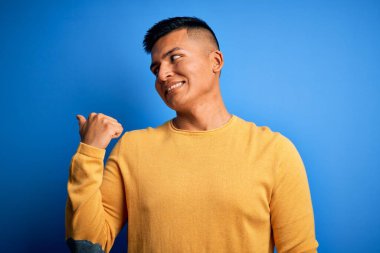 Sarı günlük süveter giyen, mavi arka planda gülümseyen, gülümseyen ve baş parmağıyla yüzünü gösteren genç, yakışıklı Latin bir adam..