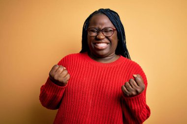 Sarı arka planda sıradan süveter giyen Afrikalı Amerikalı artı cüsseli kadın. Kolları havada ve gözleri kapalı zaferi kutlarken gülümsüyor. Kazanan konsept.