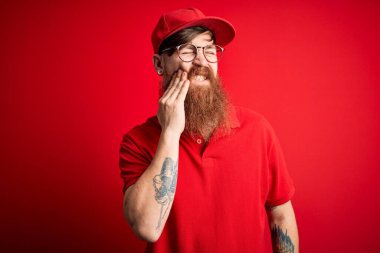 Gözlüklü ve kırmızı şapkalı genç teslimatçı diş ağrısı ya da diş hastalığından dolayı acı dolu bir ifadeyle ağıza dokunuyor. Dişçi