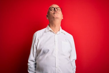 Orta yaşlı yakışıklı, beyaz gömlekli, gözlüklü, kırmızı arka plan gözlüklü, güzel ve seksi bir şekilde havada öpüşen kameraya bakan bir adam. Aşk ifadesi.