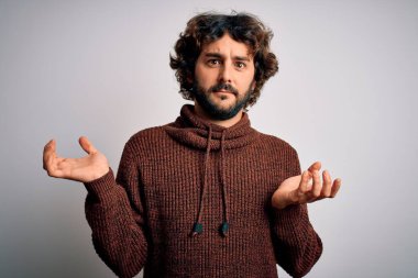 Sakallı, sıradan bir süveter giyen, beyaz arka planda duran, kafası karışmış, elleri ve kolları havada genç ve yakışıklı bir adam. Şüphe kavramı.