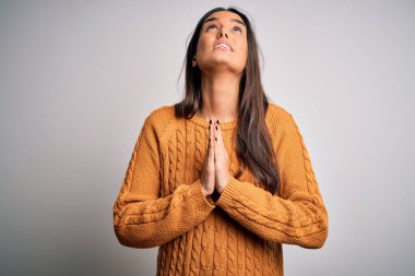 Genç, güzel esmer kadın, izole edilmiş beyaz arka plan üzerine gündelik kazak giyip elleri bağlı yalvarıyor ve dua ediyor. Yüzünde umut ifadesi var. Çok duygusal ve endişeli. Yalvarıyorum..