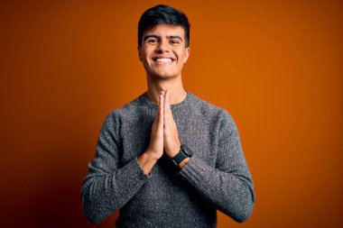 Genç ve yakışıklı bir adam, günlük süveter giyip turuncu arka planda duruyor. Elleriyle dua ediyor ve af diliyor..