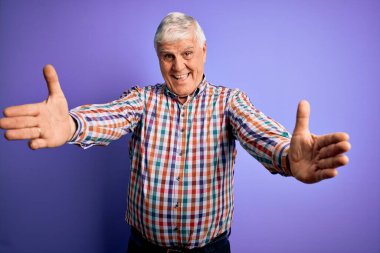 Üst düzey yakışıklı, parlak gömlekli, izole edilmiş mor arka planda sarılmak için kollarını açıp kameraya gülümseyen bir adam. Mutluluğu kucaklayan neşeli bir ifade..