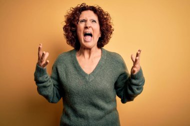 Orta yaşlı güzel kıvırcık saçlı kadın izole edilmiş sarı arka plan üzerine gündelik kazak giyiyor deli gibi bağırıyor ve agresif bir ifadeyle bağırıyor ve kollarını kaldırıyor. Hayal kırıklığı konsepti.