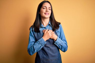 Mavi gözlü genç fırıncı kadın izole edilmiş sarı arka planda dikilmiş elleri göğsünde, gözleri kapalı ve yüzünde minnet dolu bir jestle gülümsüyor. Sağlık kavramı.