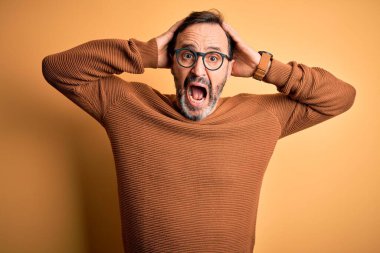 Orta yaşlı, beyaz, kahverengi süveter ve gözlüklü, izole edilmiş sarı arka plan delisi, elleri kafasında korkmuş, ağzı açık, şok geçirmekten korkan ve şaşıran bir adam.