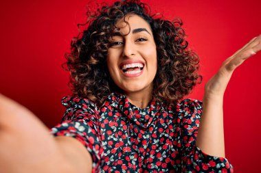 Genç, kıvırcık Arap bir kadın elbise giyiyor. Kırmızı arka planda selfie çekiyor. Çok mutlu ve heyecanlı.