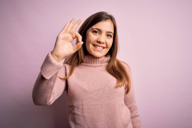 Güzel genç bir kadın pembe izole edilmiş arka planda balıkçı yaka kazak giyiyor gülümsüyor ve el ve parmaklarıyla 