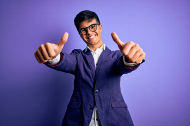 Ceket ve gözlük takan genç yakışıklı iş adamı mor arka planda olumlu bir jest yaptığını onaylıyor, gülümsüyor ve başarı için seviniyor. Kazanan jest.