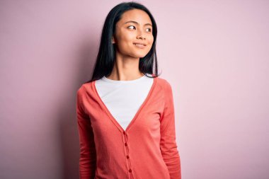 İzole edilmiş pembe arka plan üzerine sıradan süveter giyen genç, güzel Çinli bir kadın yüzünde gülümsemeyle, doğal bir ifadeyle yan yana bakıyor. Kendinden emin gülen.