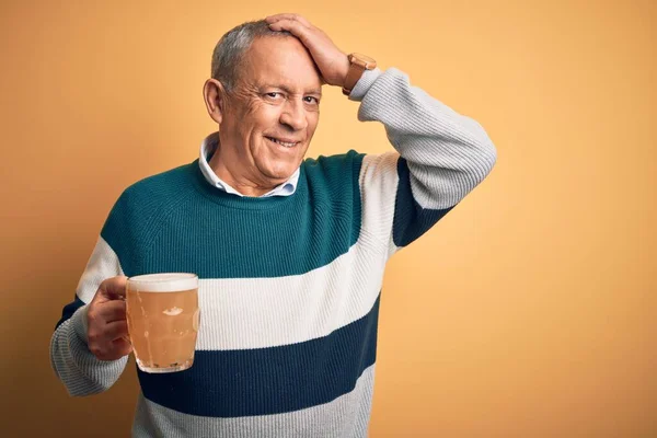 Senior handsome man drinking jar of beer standing over isolated yellow background smiling confident touching hair with hand up gesture, posing attractive and fashionable