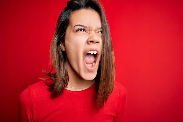 Young beautiful brunette girl wearing casual t-shirt over isolated red background angry and mad screaming frustrated and furious, shouting with anger. Rage and aggressive concept.