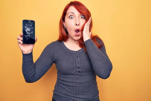 Young beautiful redhead woman holding broken smartphone showing cracked screen scared and amazed with open mouth for surprise, disbelief face