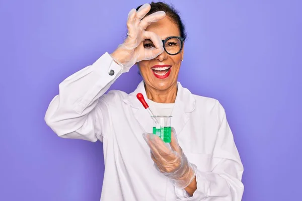Middle age senior scientist woman wearing laboratory coat holding research test tube with happy face smiling doing ok sign with hand on eye looking through fingers