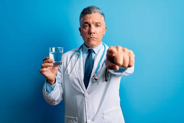 中世ハンサムな白髪の医者男飲料水のガラスの上に青の背景指差す指でカメラとあなたに 手のサイン 正と自信を持ってジェスチャーからフロント — ストック写真