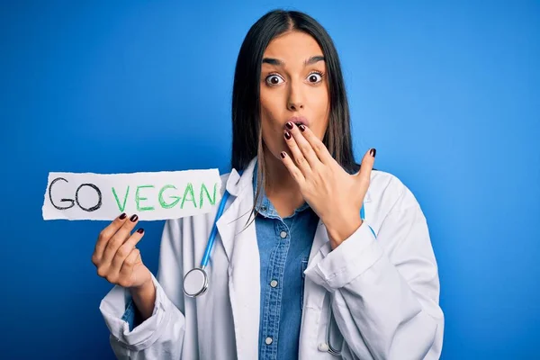 Young beautiful brunette doctor woman wearing coat holding paper with go vegan message cover mouth with hand shocked with shame for mistake, expression of fear, scared in silence, secret concept