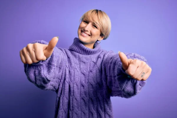 Young blonde woman with short hair wearing winter turtleneck sweater over purple background approving doing positive gesture with hand, thumbs up smiling and happy for success. Winner gesture.