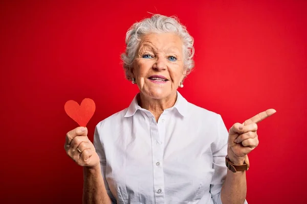 Senior beautiful woman holding paper heart standing over isolated red background very happy pointing with hand and finger to the side