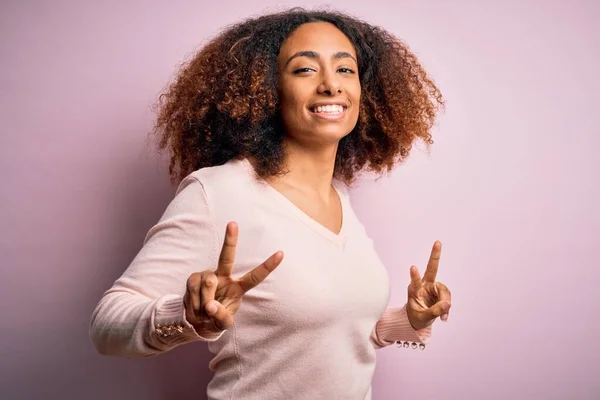 ピンクの背景にカジュアルなセーターを着たアフロの髪をした若いアフリカ系アメリカ人女性が勝利のサインをしている指を示すカメラを見て微笑んだ 第二番 — ストック写真