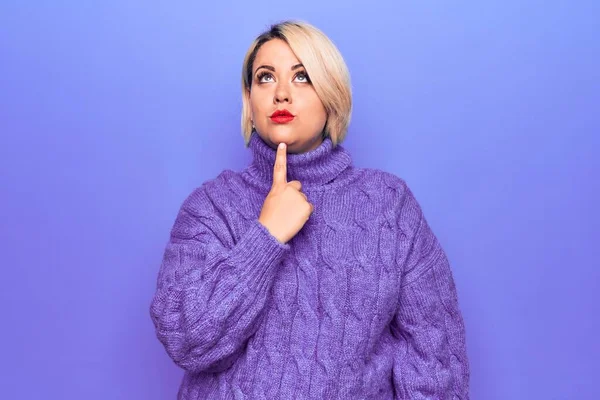 Güzel sarışın ve cüsseli bir kadın mor arka planda balıkçı yaka kazak giyiyor. Şüphe üzerine yoğunlaşmış, çenesinde parmak olan ve kafasını kaldırıp merak eden biri.