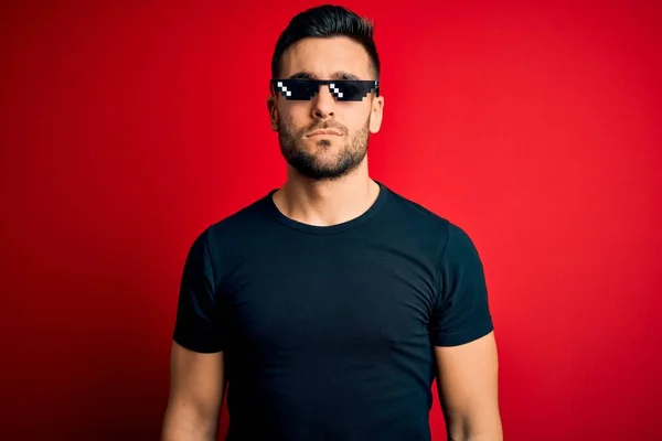 Young handsome man wearing funny thug life sunglasses over isolated red background with serious expression on face. Simple and natural looking at the camera.