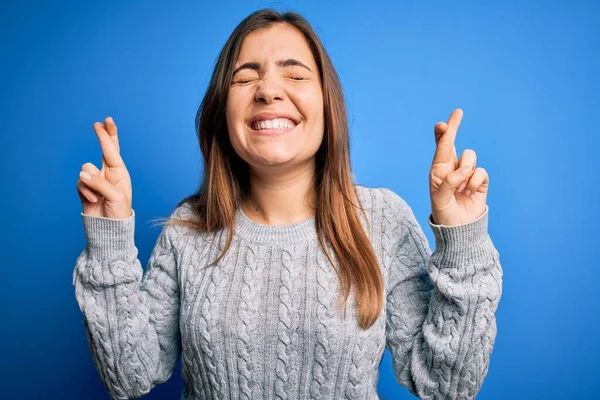 Beautiful young woman wearing casual wool sweater standing over blue isolated background gesturing finger crossed smiling with hope and eyes closed. Luck and superstitious concept.