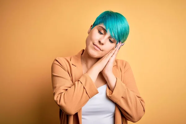 Young beautiful woman with blue fashion hair wearing casual jacket over yellow background sleeping tired dreaming and posing with hands together while smiling with closed eyes.