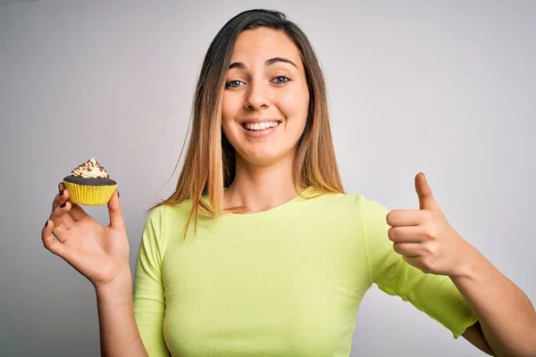 青い目の若い美しい女性食べる甘いチョコレートカップケーキ上白い背景幸せな大きな笑顔をしながらOkサイン 親指アップで指 優れたサイン — ストック写真